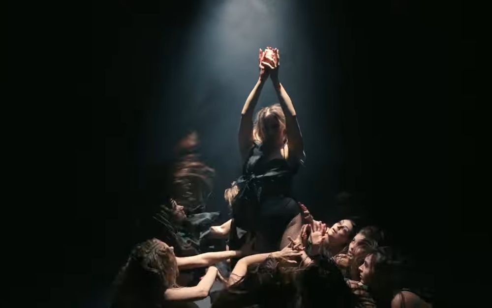 Still from Royal Dance performance, where the central dancer holds a bloody heart aloft in a spotlight, as the ensemble looks up at it