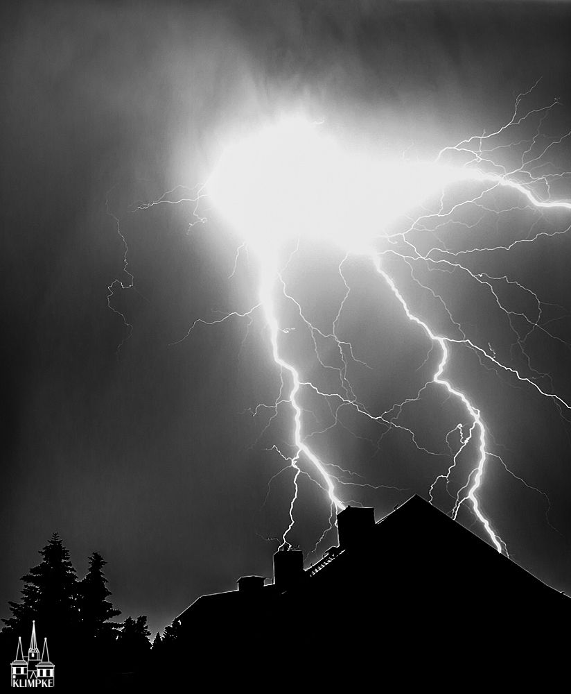 Nacht. Im Vordergrund sind die Silhouetten einen Hauses (rechts) und von Nadelbäumen (links) zu sehen.
Darüber zucken im Nachthimmel mehrere Blitze nach unten.
Ihr Licht gibt den Silhouetten eine dünne weiße Umrandung.
Schwarzweißfoto. 