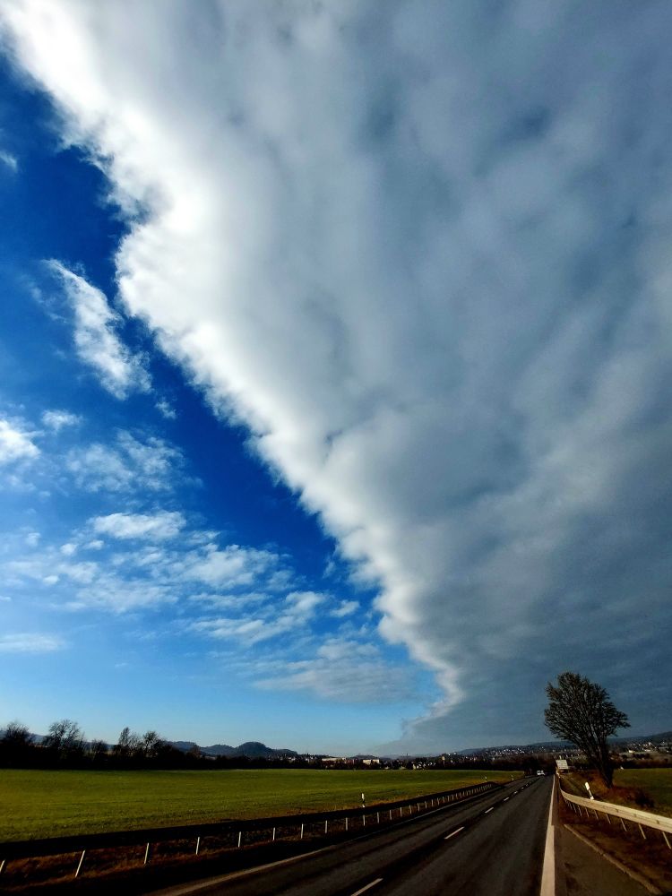 Unten ist eine zweispurige Straße, begrenze von Leitplanken zu sehen.
Darüber sind links blauer Himmel und rechts eine Wolkendecke zu sehen. Die recht scharfe Kante zwischen beiden verläuft von links oben zur Bildmitte.