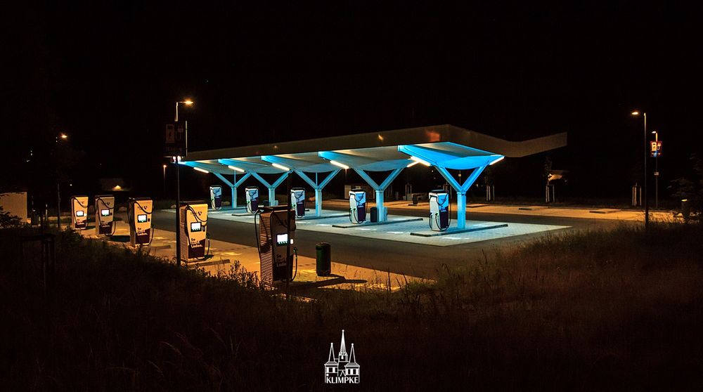 Vom Dunkel der Nacht umhüllt stehen links im Vordergrund 5 Ladesäulen in gelblichem Licht.
Dahinter stehen im Zentrum des Bildes unter einem Dach in bläulichem Licht 5 weitere Ladesäulen.