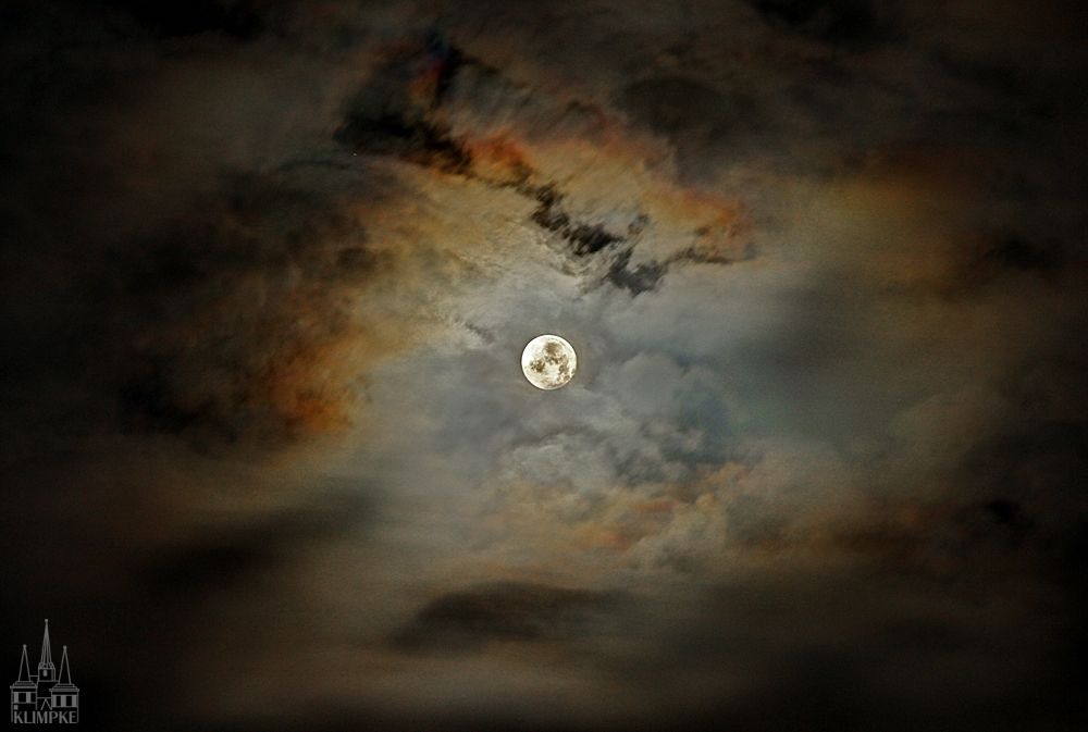 Bewölkter Nachthimmel mit dem Vollmond im Zentrum.
Das Mondlicht erzeugt in den Wolken eine farbige Corona.