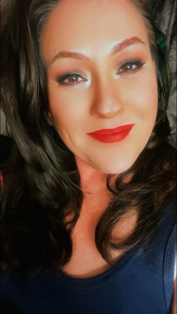 A pretty brunette with shiny long brown hair cascading over her shoulders. With classy sexy red lips stares into the camera and smiles showing a little dimple. 