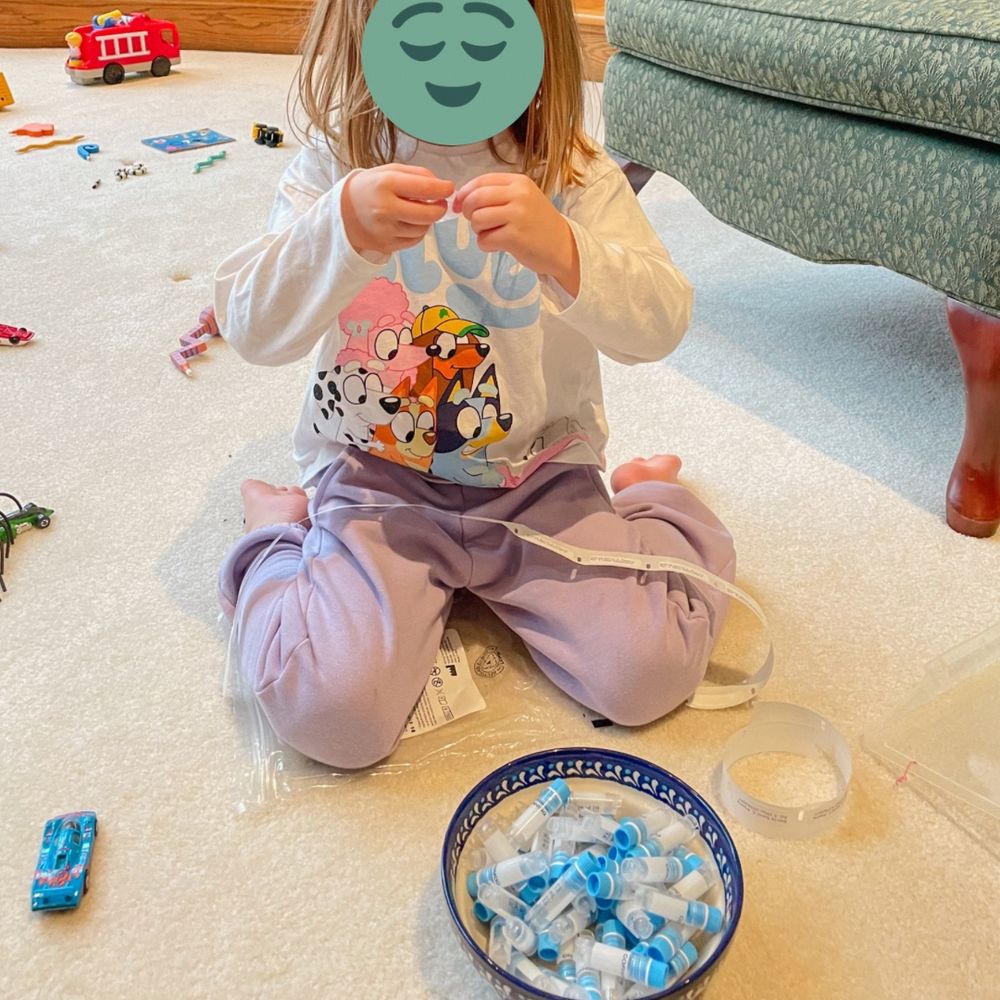 Preschooler in a bluey shirt and purple sweatpants sits on the floor labeling tubes with stickers for bio specimen collection, surrounded by toy cars