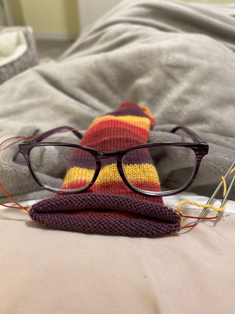 A partially knit sock in red, maroon, and yellow striped yarn lies on a blanket with the open end facing the camera. On top of the sock is an open pair of reading glasses, making it appear as though the sock has eyes and a mouth. 