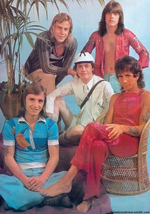 A very odd studio photograph of early AC/DC (c. 1975). This lineup features the very short-lived bass player Paul Matters (upper left with the blonde hair), who would soon be replaced by Mark Evans.

They’re all posed against a powder blue backdrop, complimented by a potted plant, a purple rug, and a rattan chair.

Singer Bon Scott is sat on the rattan chair with legs crossed, and hand on his thigh with his eyes on the camera with a subtle smirk. He’s dressed in only a very hot pink (or perhaps red?) pair of satin overalls.

Behind him (upper right), rhythm guitarist Malcolm Young is stood with his hands on his hips, facing the camera. He’s wearing jeans and a silk kimono/happi style top in a similar color to Bon’s overalls.

Lead guitarist Angus Young is sat in the middle of the group, grinning at the camera in his usual way. He’s wearing one of the very first (THE first, IIRC) iterations of his schoolboy outfit—an all white version of the iconic costume we know so well.

And finally, we arrive at the bottom left to see drummer Phil Rudd greeting us with a rare smile—albeit an awkward looking one. He’s sat on the rug with crossed legs and dressed in baby blue bell bottoms with a rather cutesy sailboat themed polo (honestly I’d like to have it) shirt. It has white elastic borders along its openings—the majority of it being a light blue color with some darker blue stripe accents. The large stripe across the chest makes up the ‘ocean’ on which the large sailboat graphic sails.

Like Phil’s shirt, the whole photoshoot seems rather uncharacteristically twee for how we normally see and envision AC/DC. They look so polite here!