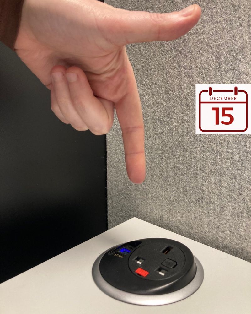 A hand points downwards towards a socket for a three-pin plug and USB charger that is fitted into the surface of a white desk.