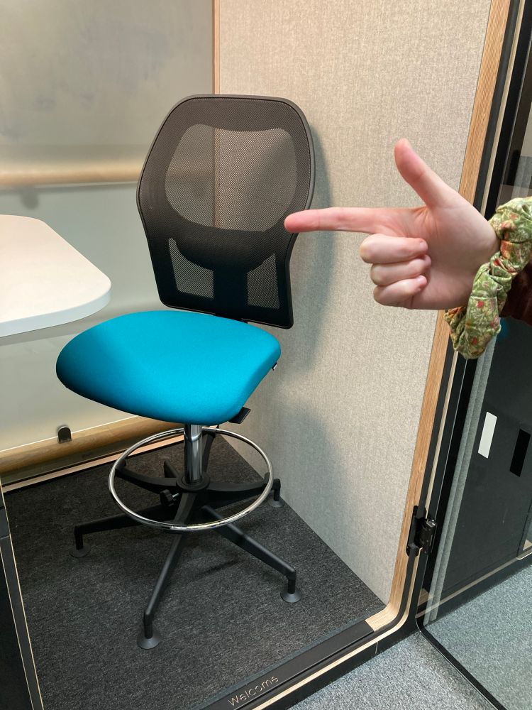 A hand points towards a height-adjustable revolving chair that stands in a study pod next to a white desk.