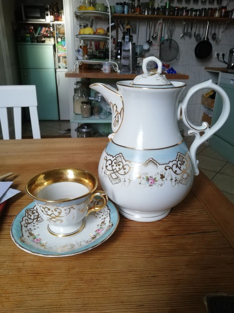 Foto von alter Kaffeekanne und -Tasse mit Gold- und Blümchenbemalung. Die Tülle der Kanne ist als Schnabel oben angesetzt.