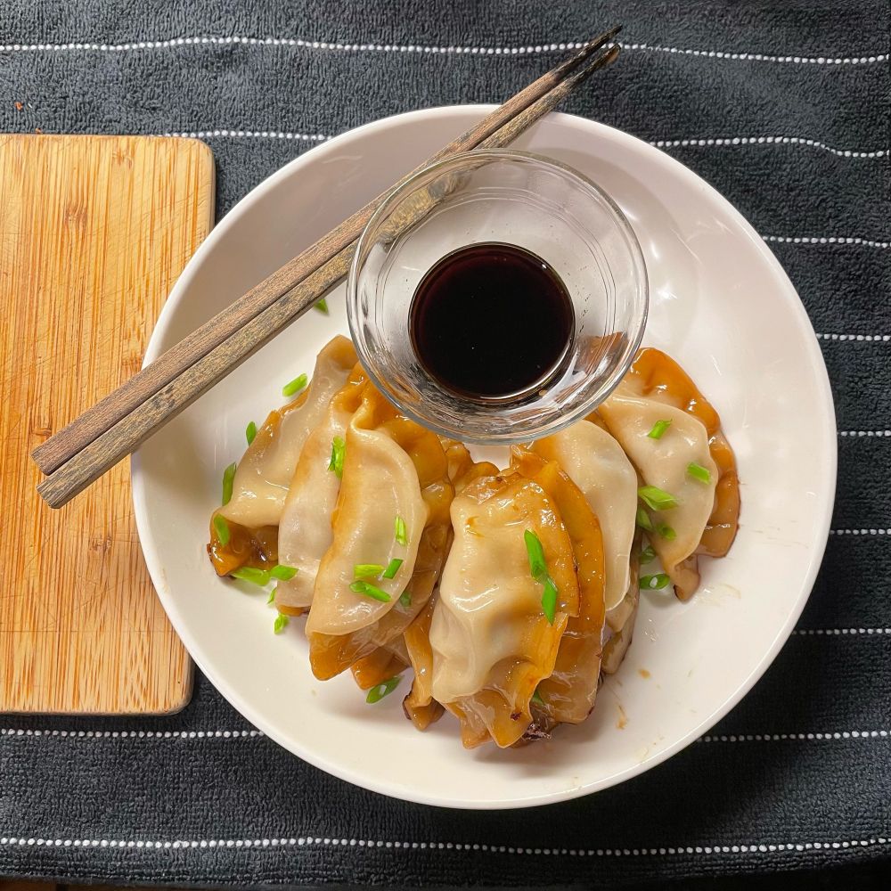 Ten gyoza neatly stacked on a white plate and topped with green onion, next to a glass dish of soy sauce and chopsticks for the devouring phase 