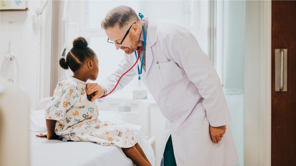 A stock photo (or possibly AI-generated) showing a physician with a stethoscope bell placed on the chest of a young girl in a pediatric patient gown. The stethoscope earpieces are clearly not in his ears. 