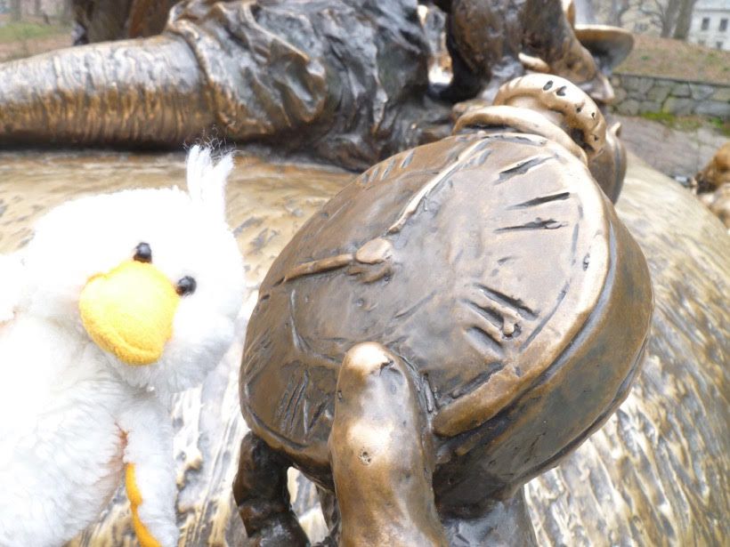 Quirk, a small, white, stuffed toy duck with a yellow beak and underwings and small, black, round eyes poses in Central Park in Manhattan with the White Rabbit’s watch from a bronze sculpture of Alice in Wonderland.