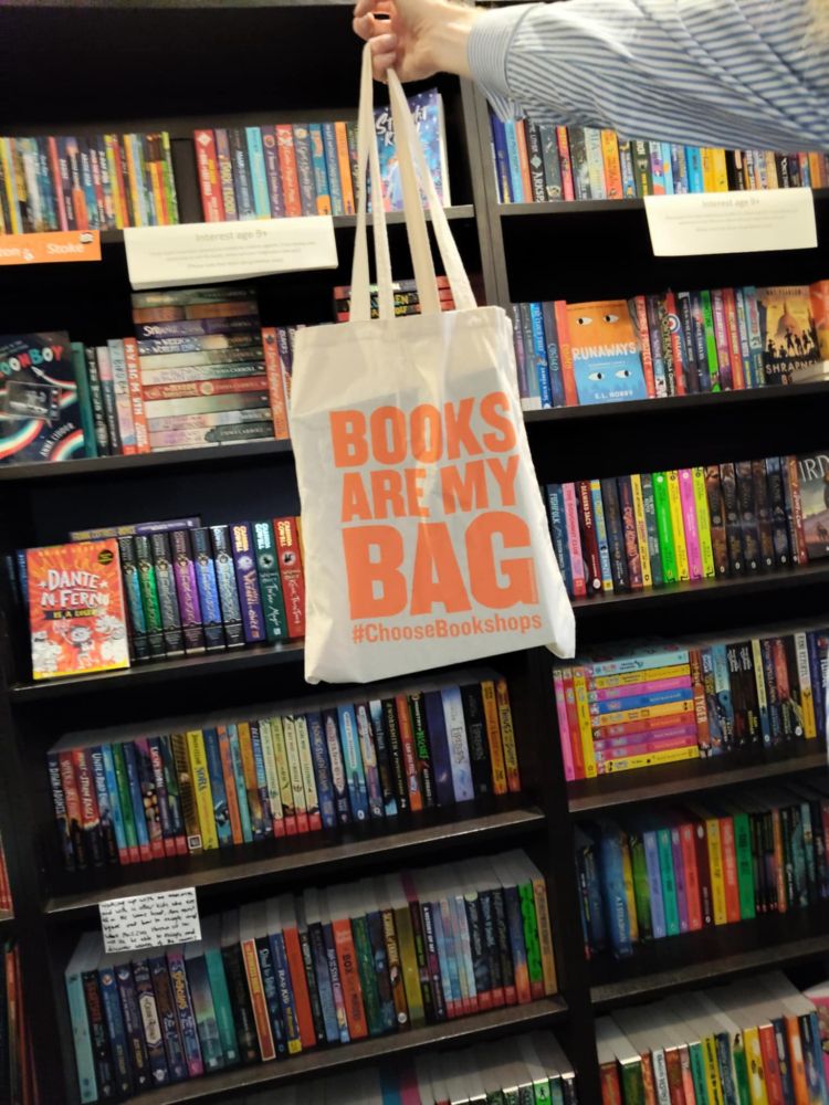 Tote bag saying Books Are My Bag being held up in front of bookcases in Wonderland Bookshop. 