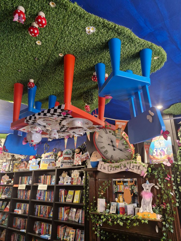 Interior of Wonderland Bookshop. A red and blue upside down tea party hangs from the ceiling and a clock hangs over the fireplace. The sign underneath reads "I'm late, I'm late" 