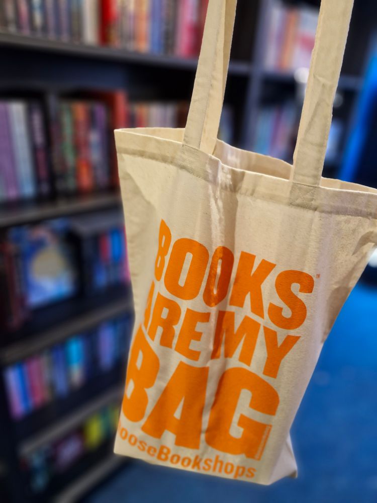 Photo of a tote bag being held up in front of bookcases. Books are my bag, choose bookshops is printed on it in orange. It is full of bookish goodies. 