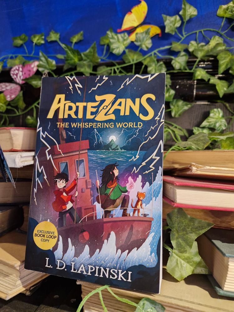 Proof copy of Artezans the Whispering World by LD Lapinski displayed on a pile of old books in Wonderland Bookshop 