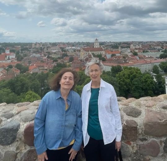 Two women stand in front of a stone wall at a scenic overlook, with a panoramic view of Vilnius, Lithuania in the background. The cityscape features red-tiled roofs, church towers, and clusters of trees under a cloudy sky. The woman on the left wears a blue jacket, and the woman on the right wears a white jacket over a teal shirt.