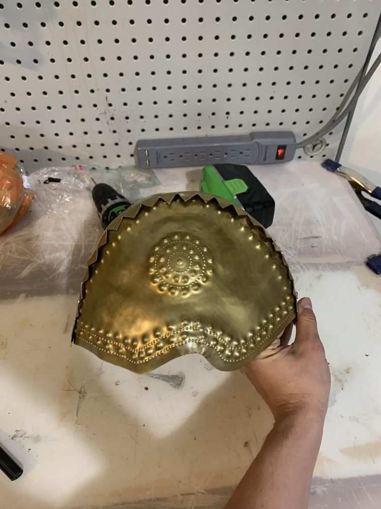 A hand holding another view of the same gold-colored armor piece, showing decorative circular rivet patterns and scalloped edging. The workbench and pegboard wall with tools are visible in the background.