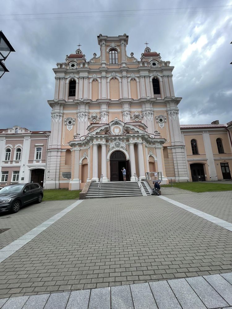 Front view of a grand Baroque-style church with peach and white tones, ornate columns, and sculpted details. The building features twin towers with domes and a central pediment above the main entrance. A person stands at the open door at the top of a staircase, while another sits nearby on the right. The sky above is cloudy and dramatic.