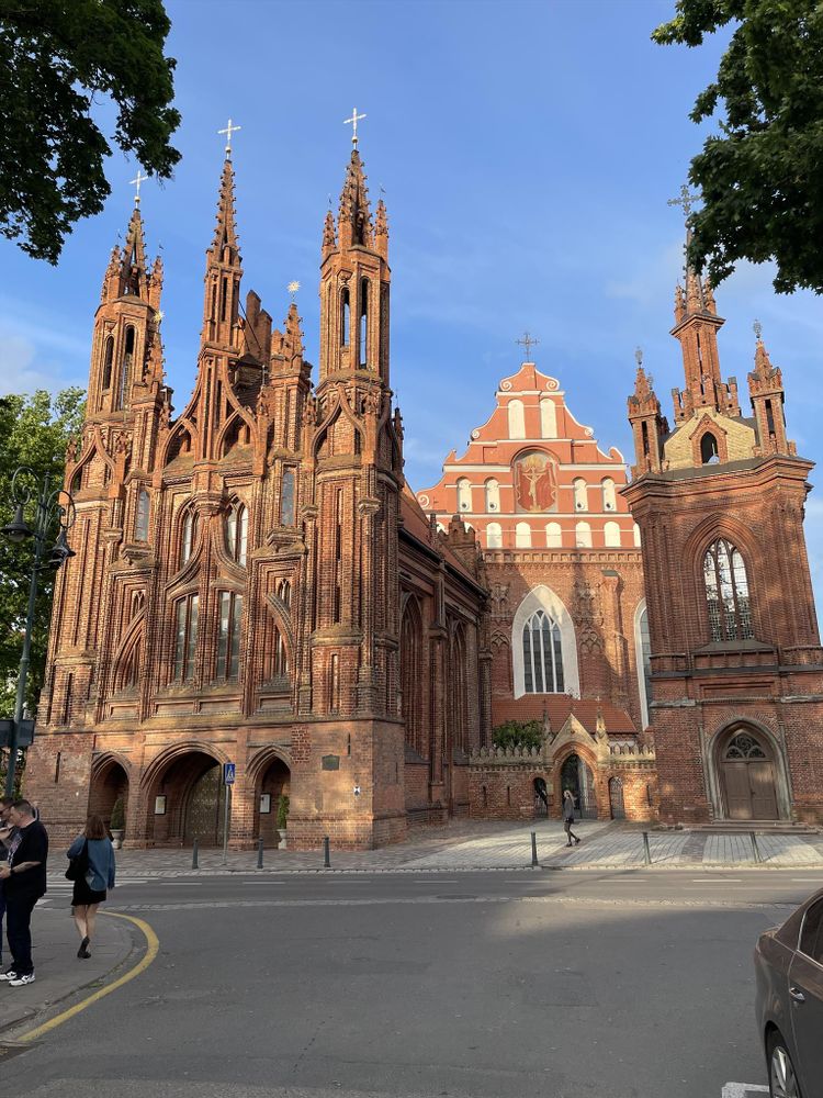Gothic-style church built from red brick, featuring tall, narrow spires with crosses and intricate detailing. The facade includes pointed arches and vertical ornamentation. Behind it stands a larger red-and-white church with a stepped gable and arched windows. Several people walk on the street and sidewalk in front, with trees framing the scene and a clear blue sky above.