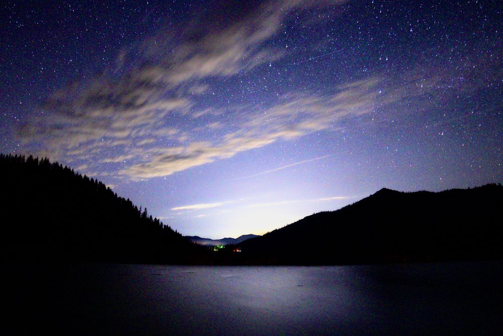 A photo of the night sky with light cloud cover and stars over a lake with the silhouette of nearby mountains on the left and right sides of the horizon. There’s a glow on the center of the horizon between the mountains shining light over the lake.