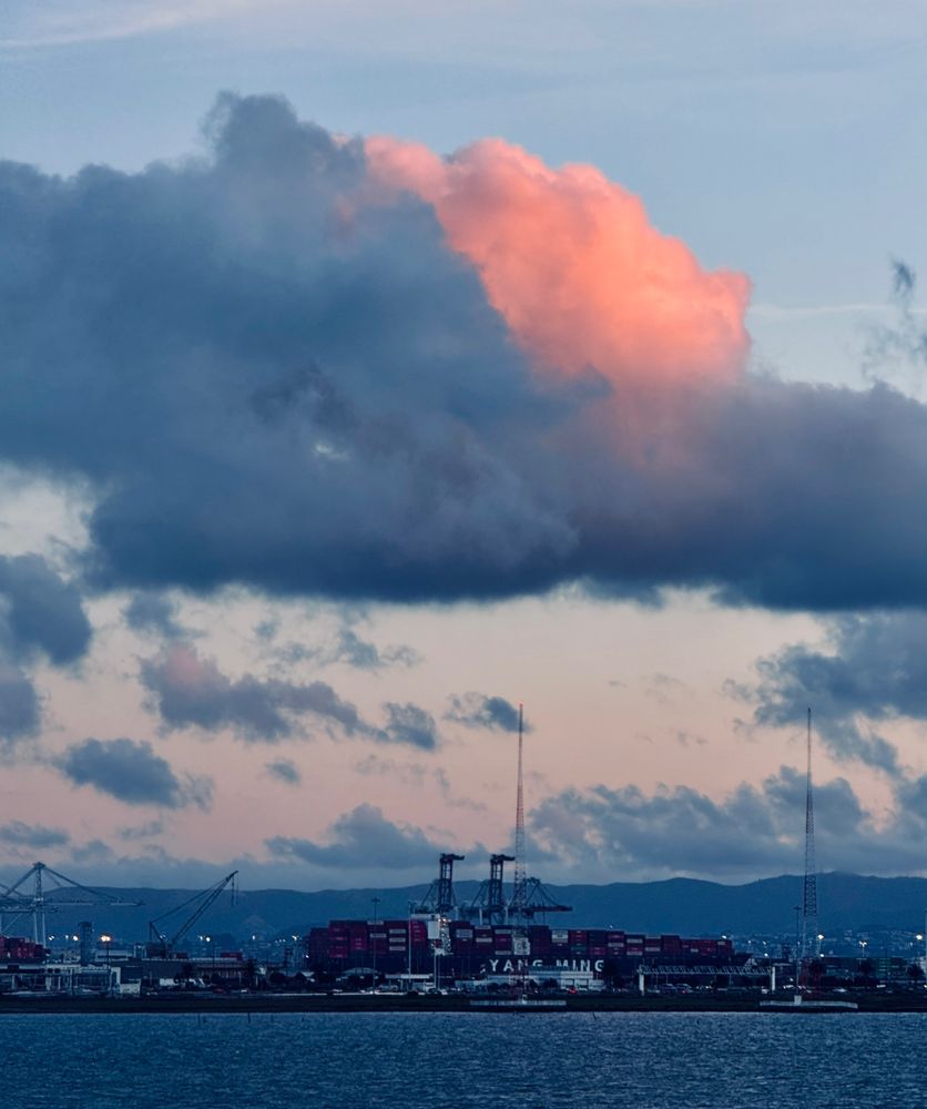 Dark bluish-gray and pink clouds over Oakland, California. Port of Oakland is in background and water is dark blue.