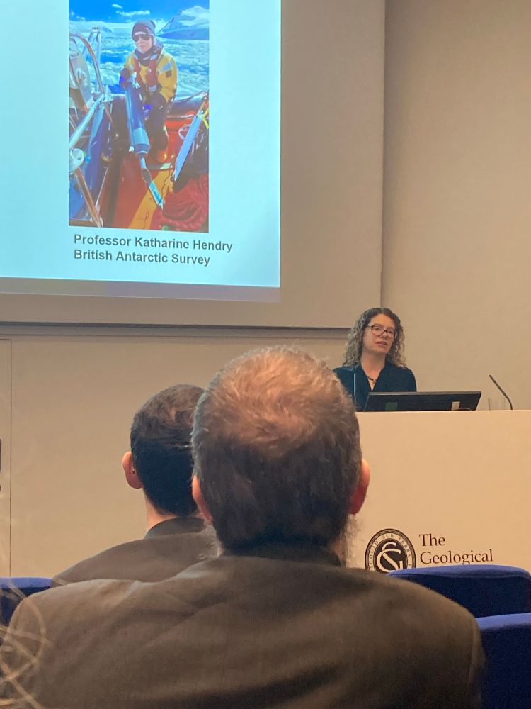 Photograph of Prof Kate Hendry, British Antarctic Survey, accepting the Bigsby Medal, in front of an image of Kate on a small boat on Antarctica