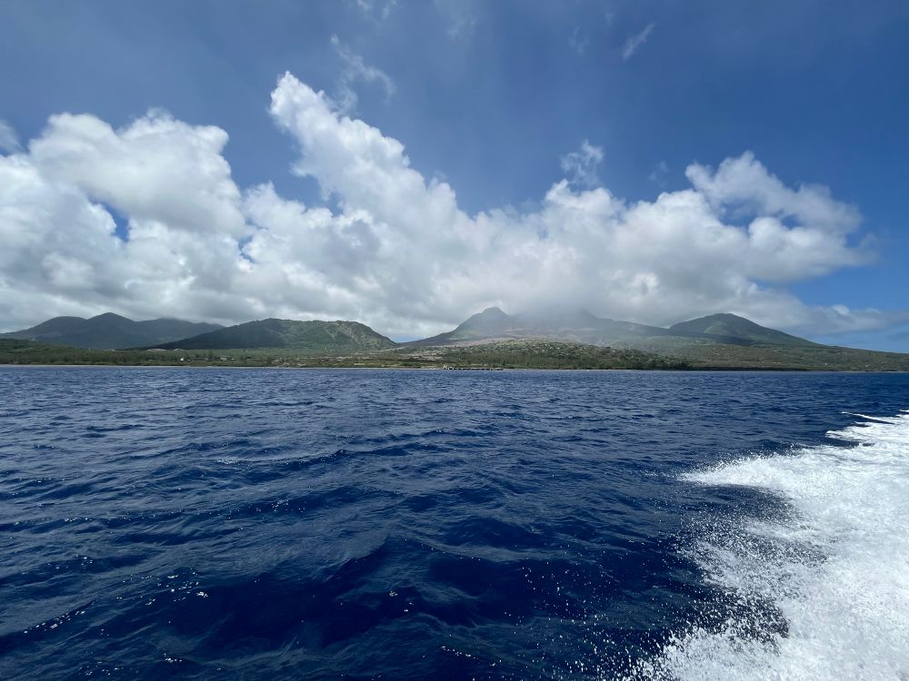 Photographic view across the sea of southern Montserrat, with the hazy green hill of the Soufriere Hills Volcano in the background, blue sky and clouds above. 