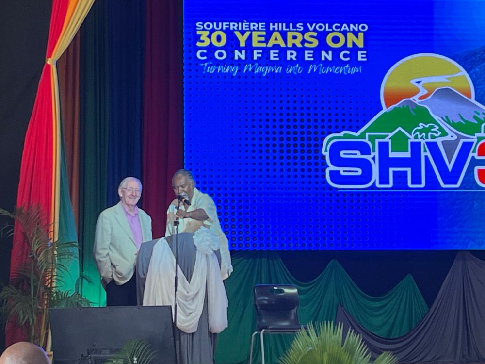 Photograph of Frank Savage in a pink shirt and linen jacket, and the Honourable Reuben T Meade, adjusting the microphone, on a stage in front of the SHV30 banner. Savage explains ‘my business suit is still on holiday in Antigua’.