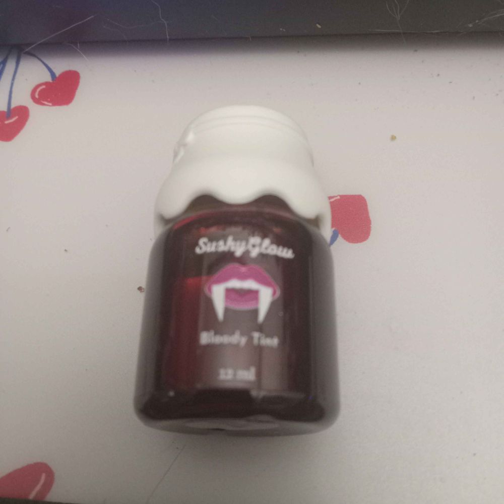 A bottle of Bloody Lip Tint from Sushy Glow Cosmetics - in the shade Bleeding Crystal. The jar is stout with a jam-jar like plastic white cap. There is a picture of a mouth with dark pink lips and exaggerated long white fangs. It is a 12ml bottle. The item is laid out on a light pink mat with red heart-shaped cherries and greenish-blue stems, some stems have one leaf on them while others don't.