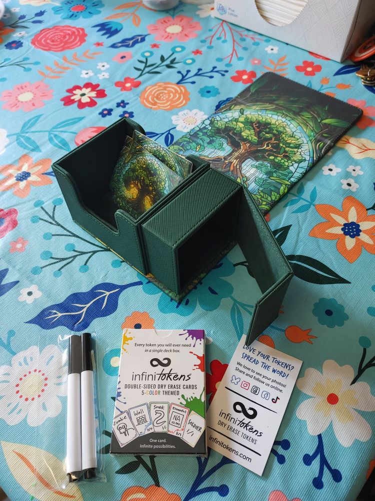 Picture of a dark green box for holding cards, unfolded with an image of a stained glass tree on the inside, and a smaller secondary box attached on the side. In front of the box is a park of multicolored Infinitokens, which are dry erase reusable cards for tabletop gaming, and a pair of markers for the tokens.