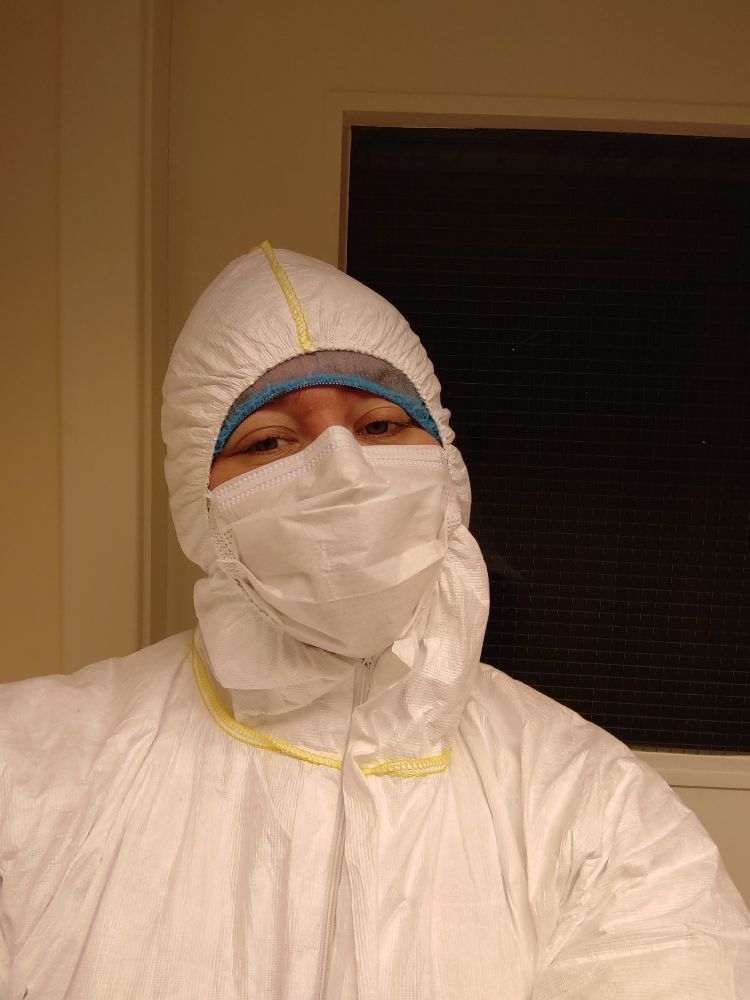 Lucy is her aDNA lab gear. She is wearing a white hazmat with a hood, a blue hairnet just visible, and a mask. You can tell that she is smiling (just!) from her eyes. She is in front of a door which leads into the lab.