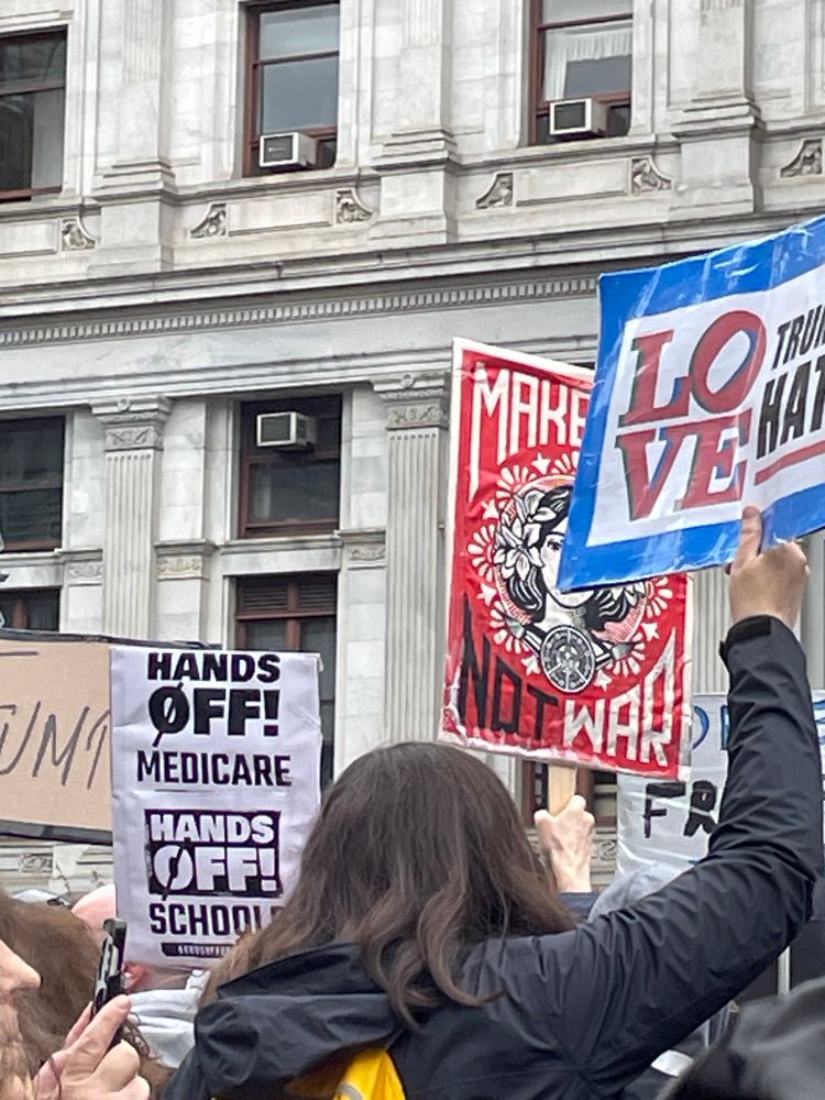 Rally with signs saying (1) “Hands Off Medicare; Hands Off Schools”. There are two other signs but words are cut off. 