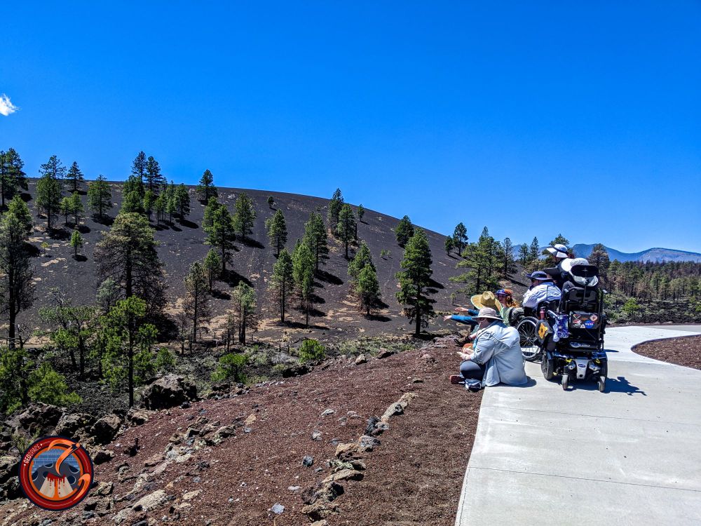 A group of students and faculty, sitting on a sidewalk and in wheelchairs, look out over the red and black cinder hills of Sunset Crater National Monument.