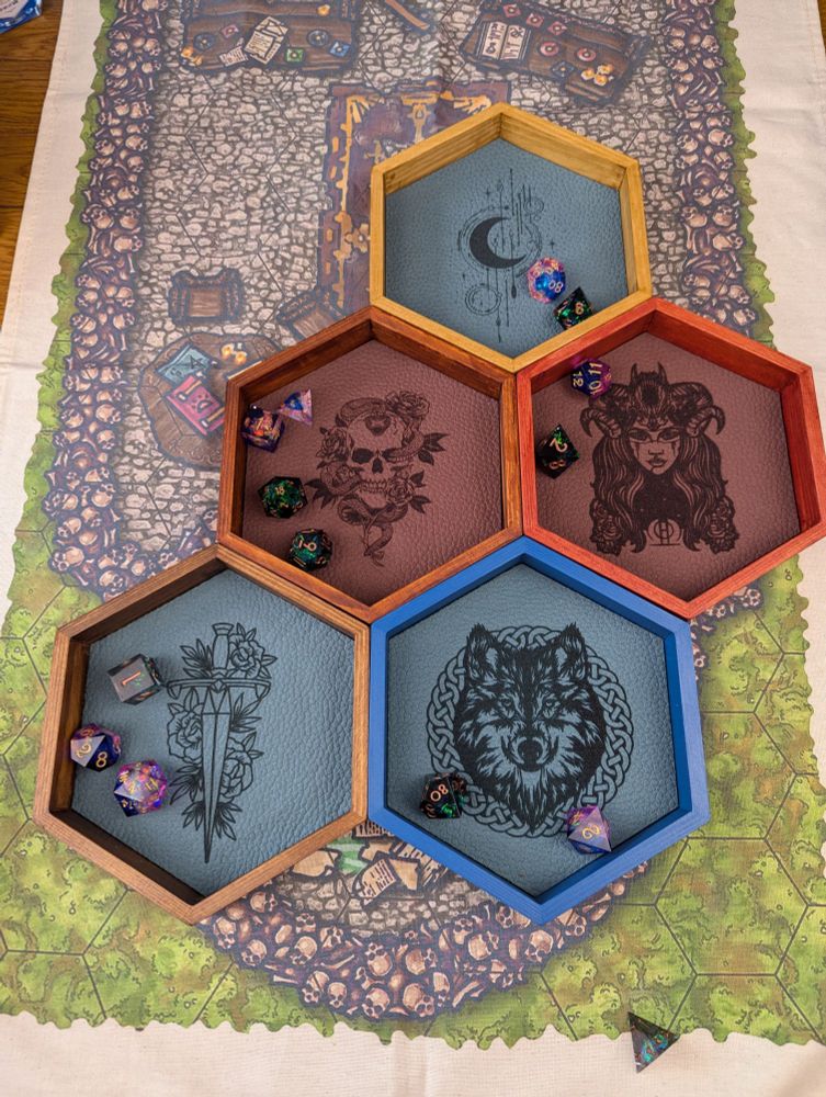 5 hexagonal wooden dice trays. Each one is a different colour - blue, oak, red, dark rosewood and country walnut. The bases are engraved with a sword and flowers, a Celtic wolf, a skull and snake, a horned woman, and a moon and stars motif 