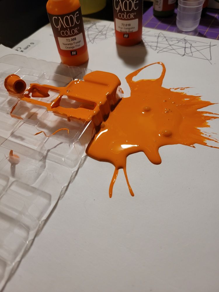 Mein Schreibtisch mit weißem Untergrund, auf dem ein Teil einer Mischpalette zu sehen ist, die orangene Farbe ist mittig fast komplett über den Untergrund und die Mischpalette gelaufen,weil der Dosierverschluss abgefallen ist. Im Hintergrund sind zwei Flaschen der Farbe.