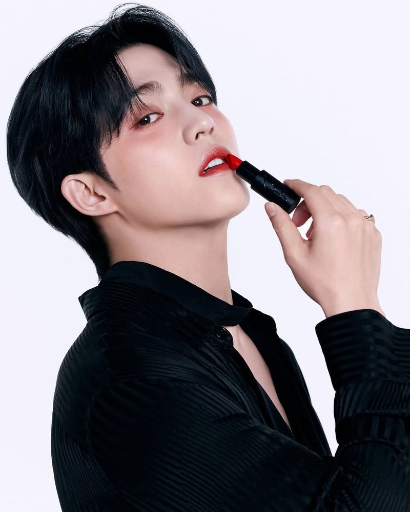 scoups applying red lipstick, he's wearing a black longsleeve shirt with a chest cutout
