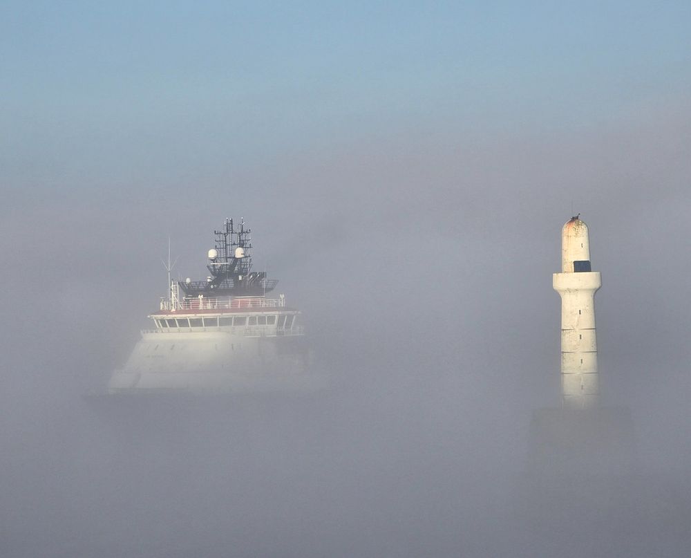 A ship appears out of a bank of fog. Only the top half is visible. To the right is the beacon at the end of the harbour breakwater.