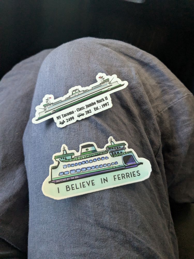 Two stickers shaped like ferries on a gray pant leg.
Top sticker is long and thin and realistic in style. On the bottom it says MV Tacoma • Class: Jimbo Mark II
👥 2499 🚗 Est.: 1997

Bottom ferry sticker is drawing style shorter And happy like it has a face and bulbous with two rows of windows
It's designed by " Sketches by the Sea" and says "I believe in Ferries"

