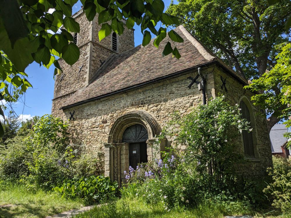 A small stone-built Norman church with arched doorway to the nave and a tower. It is a sunny day and there are spring flowers by the door.