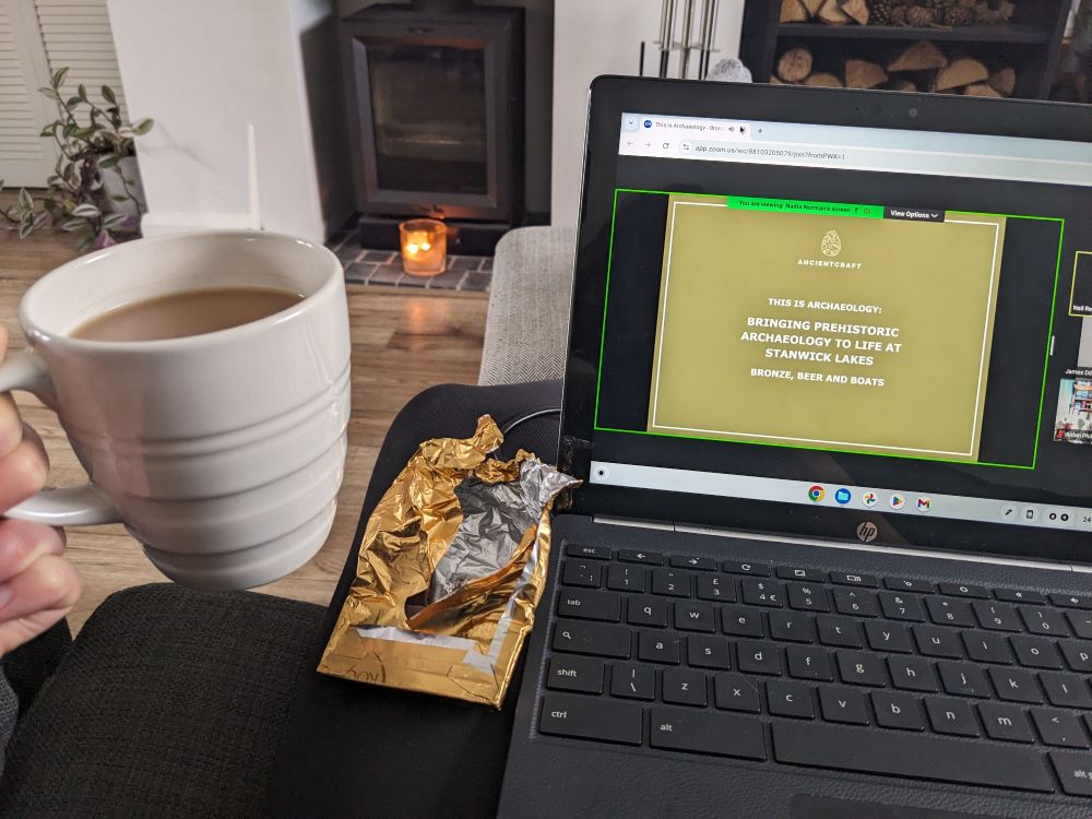 A hand holds a cup of tea in a cream mug on the left, with a laptop showing the opening slide of a 'This is Archaeology' talk on the right. In the middle is an open chocolate bar on a lap  and in the distance is a lit candle in front of a log burner.