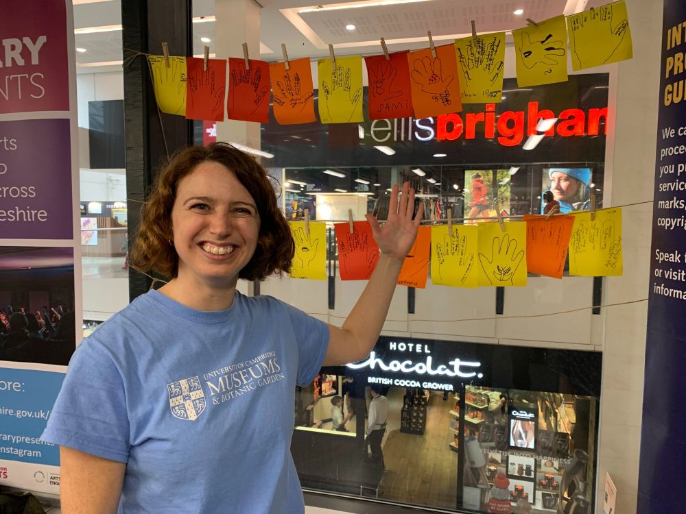 A woman wears a blue t-shirt with a University of Cambridge Museums logo, and holds up her hand in front of a display of drawn hands on paper, hanging up in front of the window of a shopping centre.