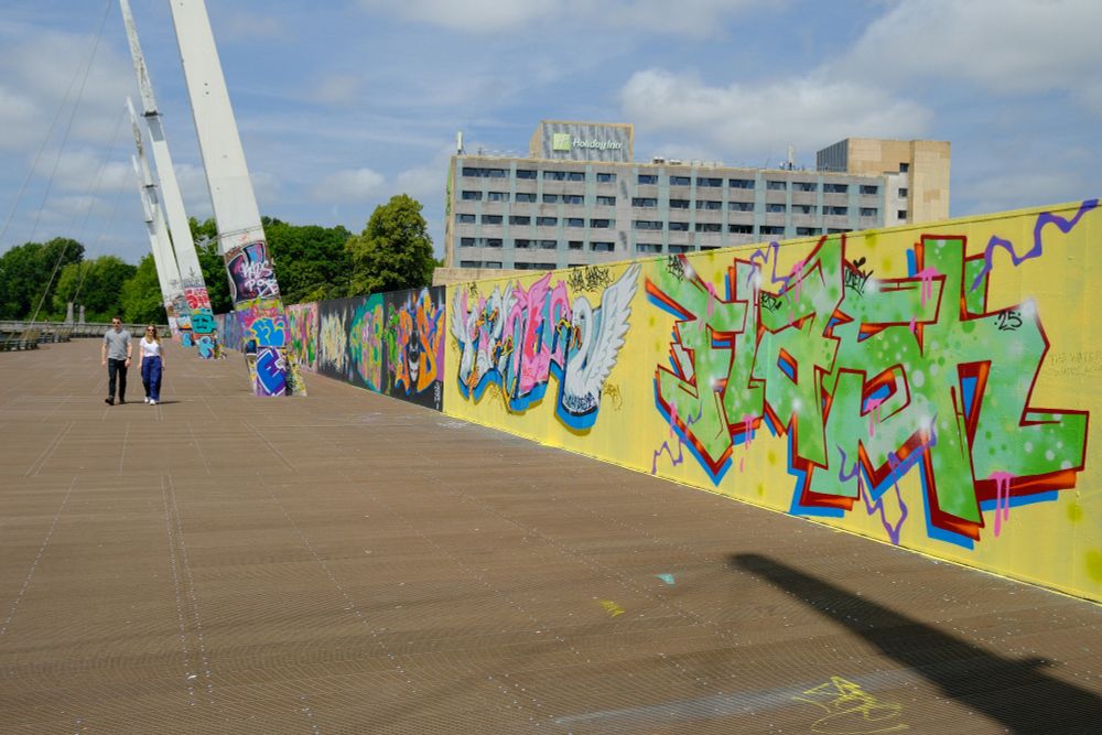 A photograph of a wall containing a variety of different graffiti art pieces located on the Millennium Walk near the Principality Stadium and it's adjacent Holiday Inn on a peaceful, partially cloudy afternoon.