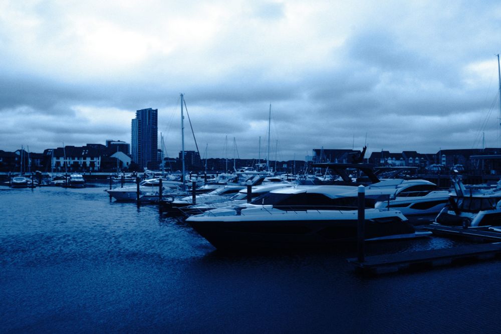 A landscape photograph featuring various yachts and boats at various distances parked harbour in Southampton near a residential area, on a dim, cloudy afternoon.