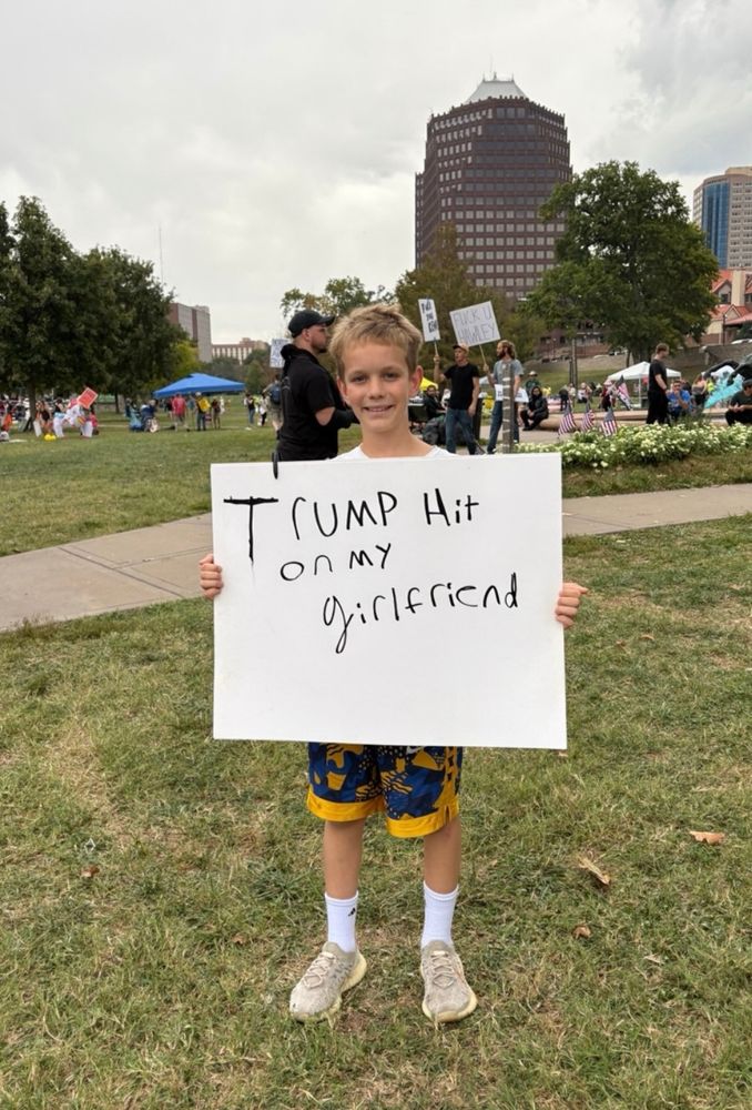 Young boy holding a sign at a protest that reads "Trump hit on my girlfriend".