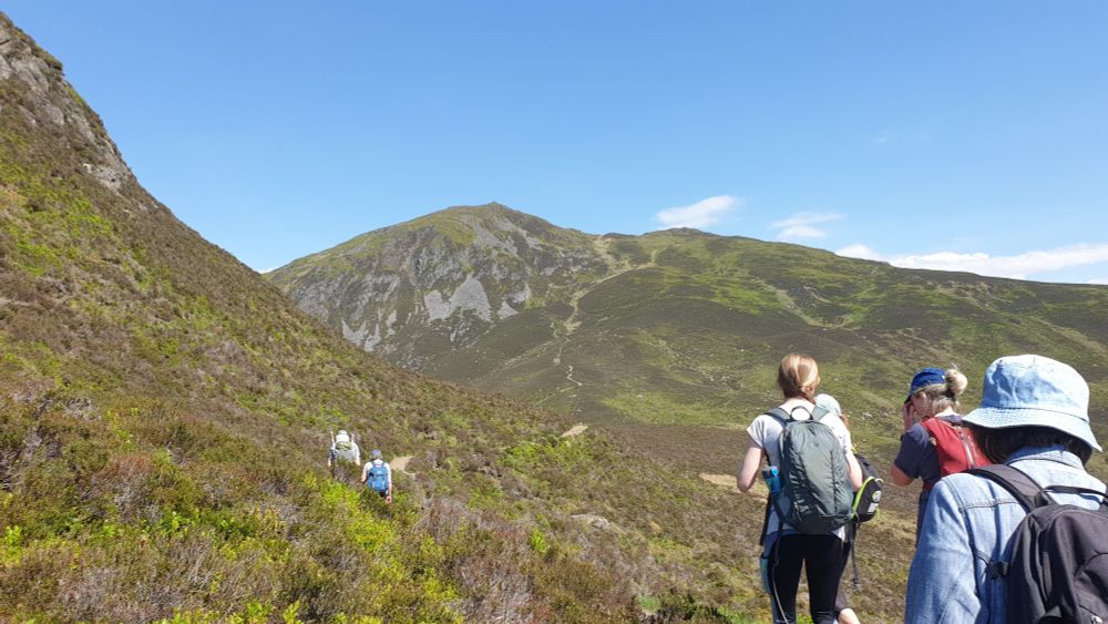 The green and heather-covered slopes of the Scottish Ben Vrackie hill seen from the path, with the backs of three people walking towards it on a sunny day with clear skies.