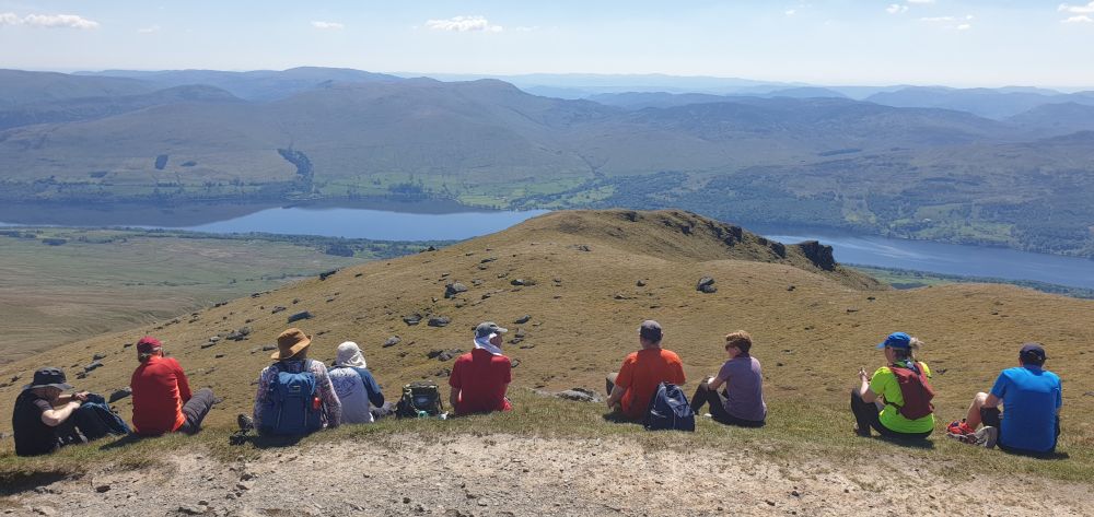 Line of hillwalkers seated on top of a mountain overlooking the blue waters of a loch and lower hills in the distance.