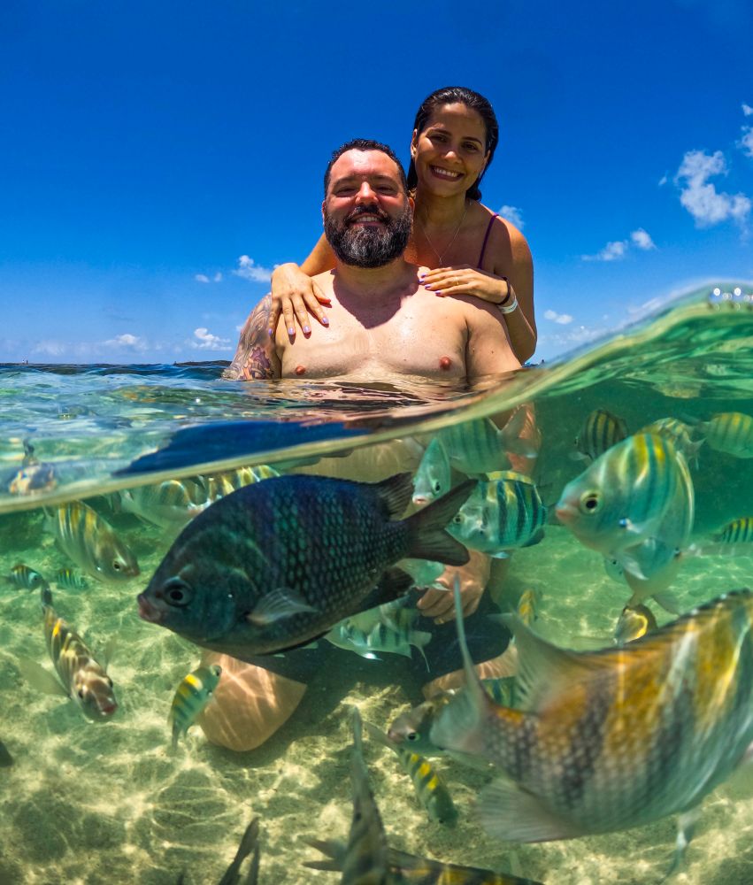 Eu, morena de cabelos castanhos e biquíni roxo, atrás do meu marido, homem branco de barba grisalha e cabelo escuro, que está sentado nas piscinas naturais de Maragogi, com vários peixinhos na frente da foto e um céu azul