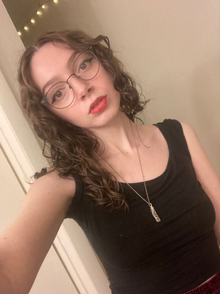 A selfie of me (Serana) my medium length brown curly hair is down and I’m wearing winged eyeliner and lip tint. I have a black top with a small clear quartz necklace. You can see a little bit of the top of my velvet purple skirt.  
