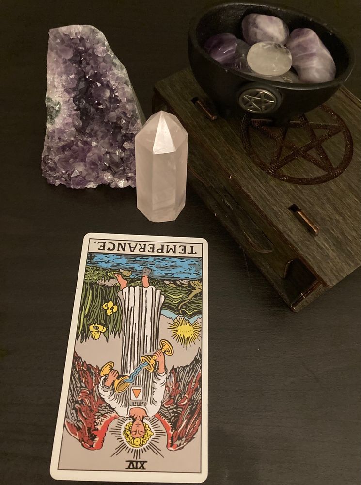 A photo of the card Temperance reversed next to the tarots container (a black box with a pentagram) and a mini Calderon with some clear quarts/amethyst inside it. There is an also a large amethyst and rose quartz next to the box. 