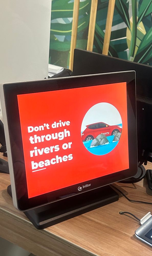 A picture of a computer monitor at a desk at a rental car agency. The monitor displays a car going through rocky water and a message reads, “Don’t drive through rivers or beaches.”